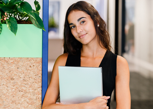 Uni student holding a laptop and leaning on shelf with indoor plant