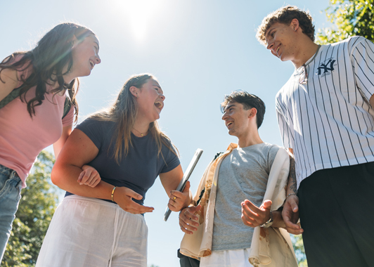 Four students standing together and laughing outside under a sunny blue sky