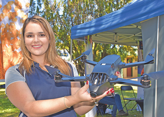 profile image of Dayna Scapin holding a drone with a marquee and trees in the background. Photo taken by Rhys Thomas