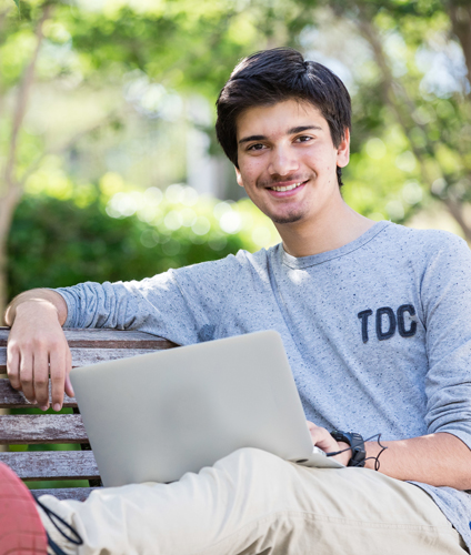 Guy with laptop reclining with one arm resting and legs up on a park bench, smiling to camera